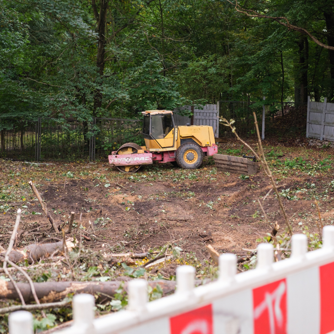Bagger steht auf einer erdigen Fläche. Im Vordergrund ist ein Bauzaun zu sehen.